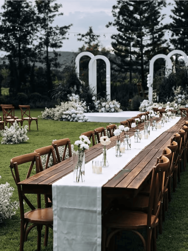Outdoor wedding setup with long wooden table and white floral arches